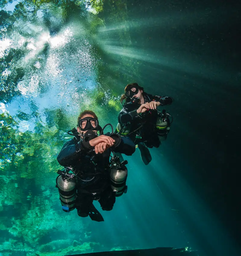 Two scuba divers exploring a cenote underwater, illuminated by dramatic sunbeams piercing through the clear turquoise water