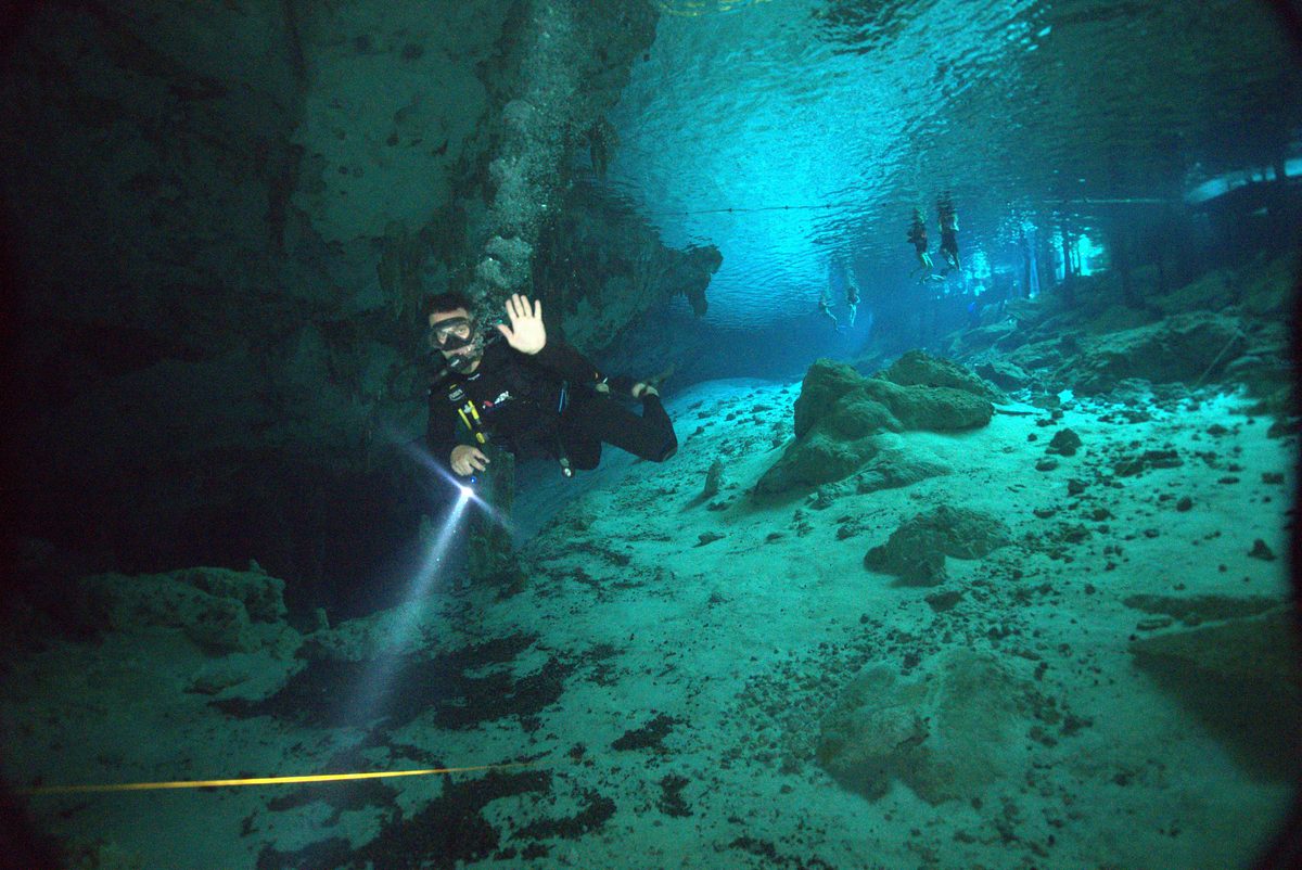 Buzo saludando durante el buceo cenote Dos Ojos mientras otros buceadores entran a la caverna al fondo