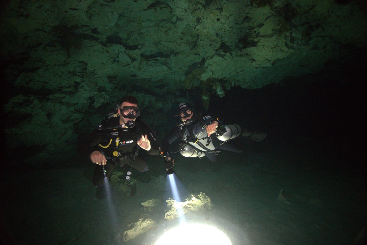 Dos buzos técnicos iluminando formaciones rocosas dentro de una caverna oscura en Dos Ojos